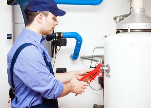 A man in a plumber's work uniform and blue baseball cap holding a clasped wrench to a valve as if tightening it.
