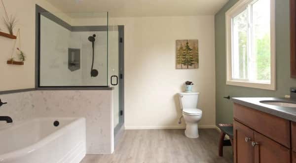 A bathroom with a glass shower, white tub, white toilet, a large two-paned window on the right wall, and a sink with wooden doors.